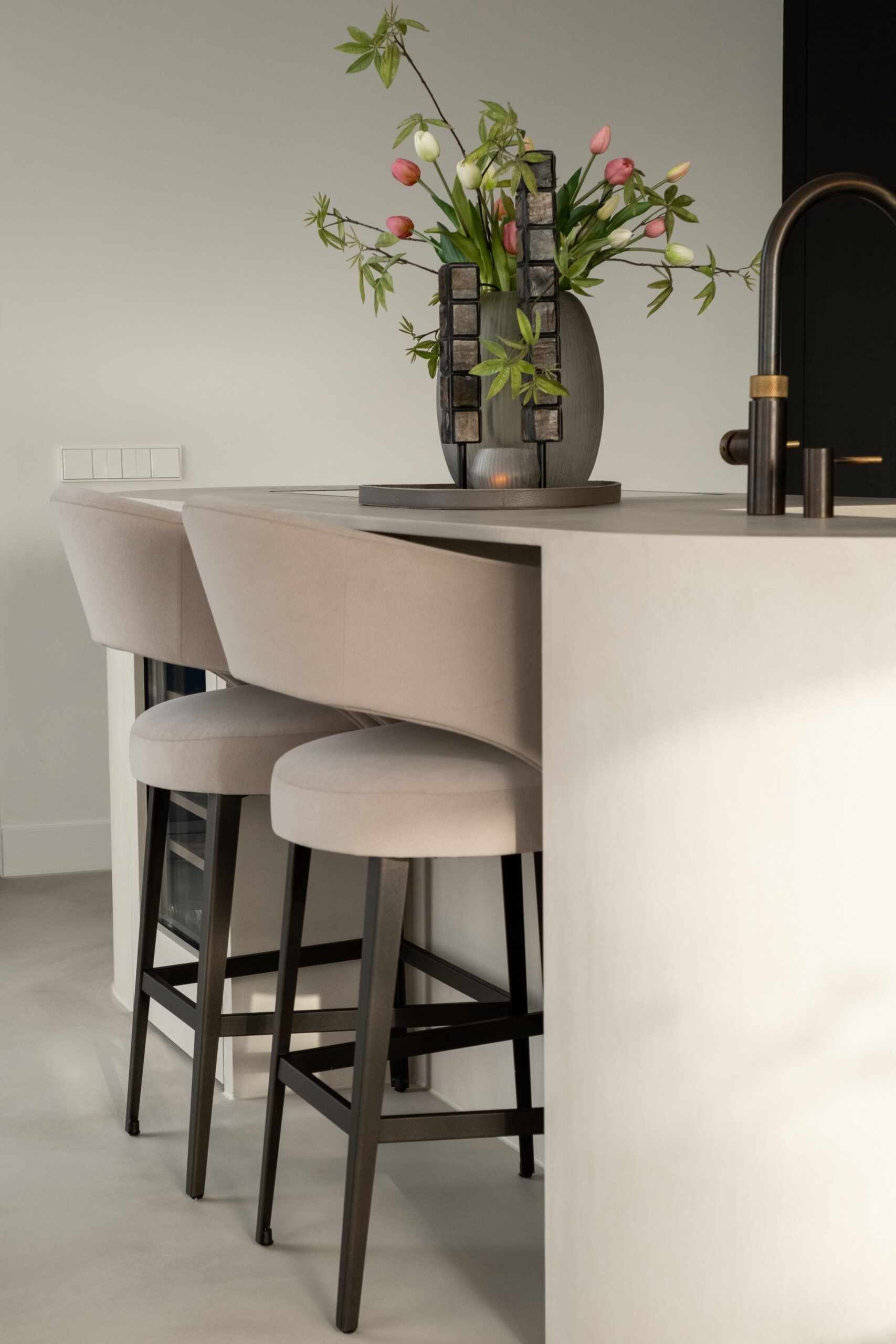 Sfeervol detail van beige gestoffeerde barstoelen aan een strak kookeiland met bloemen en een bronskleurige kraan in woning Uddel door Kardol Hout- en Interieurbouw