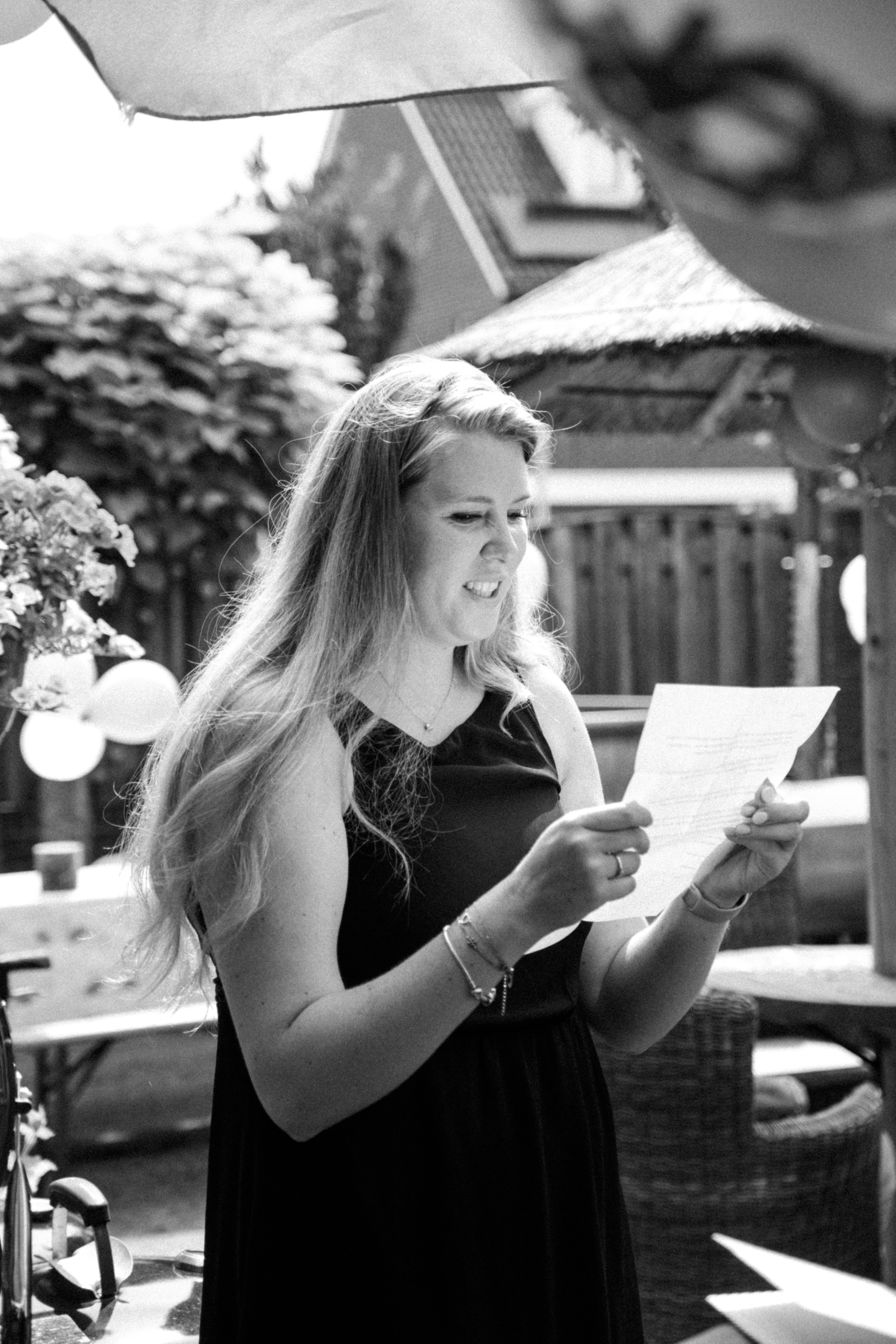 Zwart-wit foto van een lachende vrouw die een brief voorleest in de tuin tijdens de bachelorette party van Anne