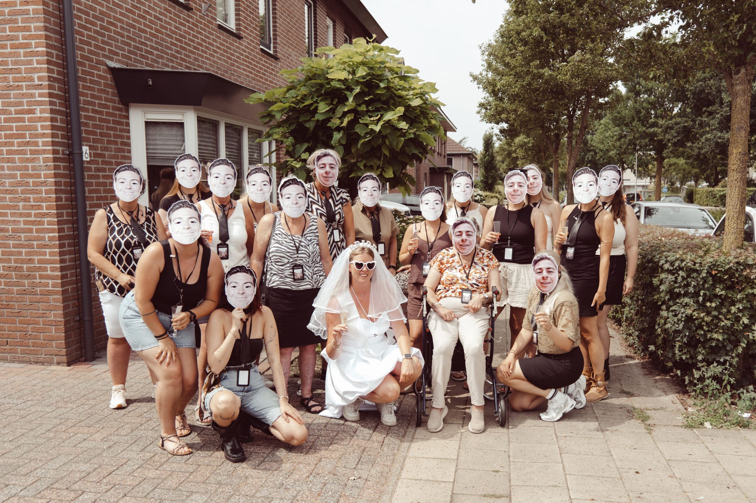 Groep vrouwen met geprinte gezichtsmaskers op poseert op straat rondom de bruid in een witte jurk tijdens de bachelorette party van Anne
