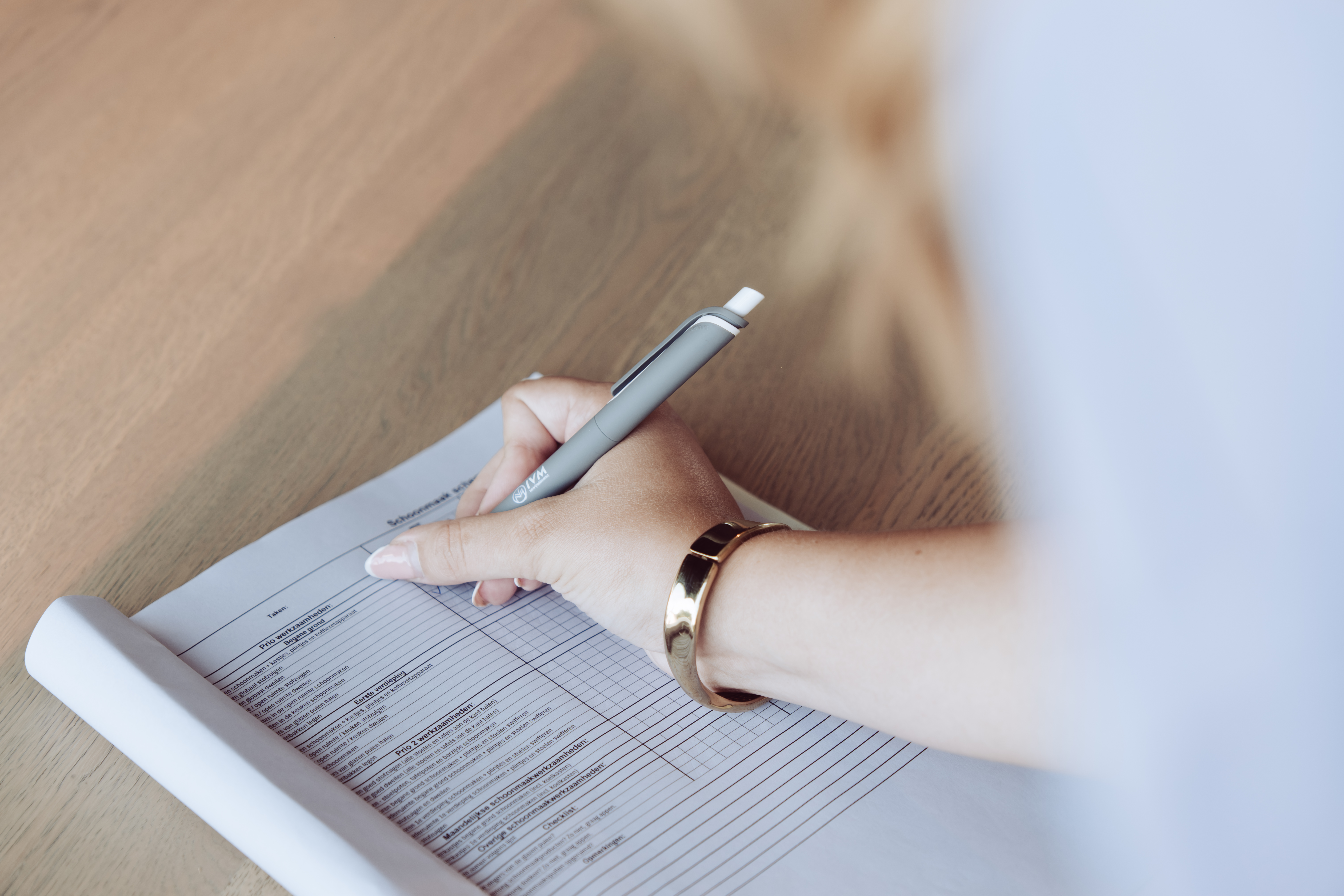 Close-up van een medewerker die met een IVM pen een schoonmaak- en takenlijst invult op een houten tafel bij IVM Bedrijfsdiensten Lunteren
