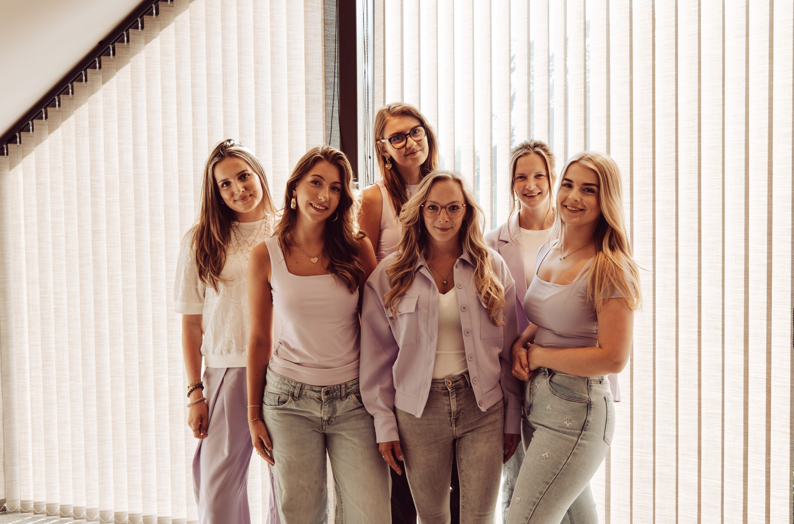 Groepsfoto van het vrouwelijke team van IVM Bedrijfsdiensten Lunteren in stijlvolle lichtpaarse en witte kleding voor een raam met jaloezieën