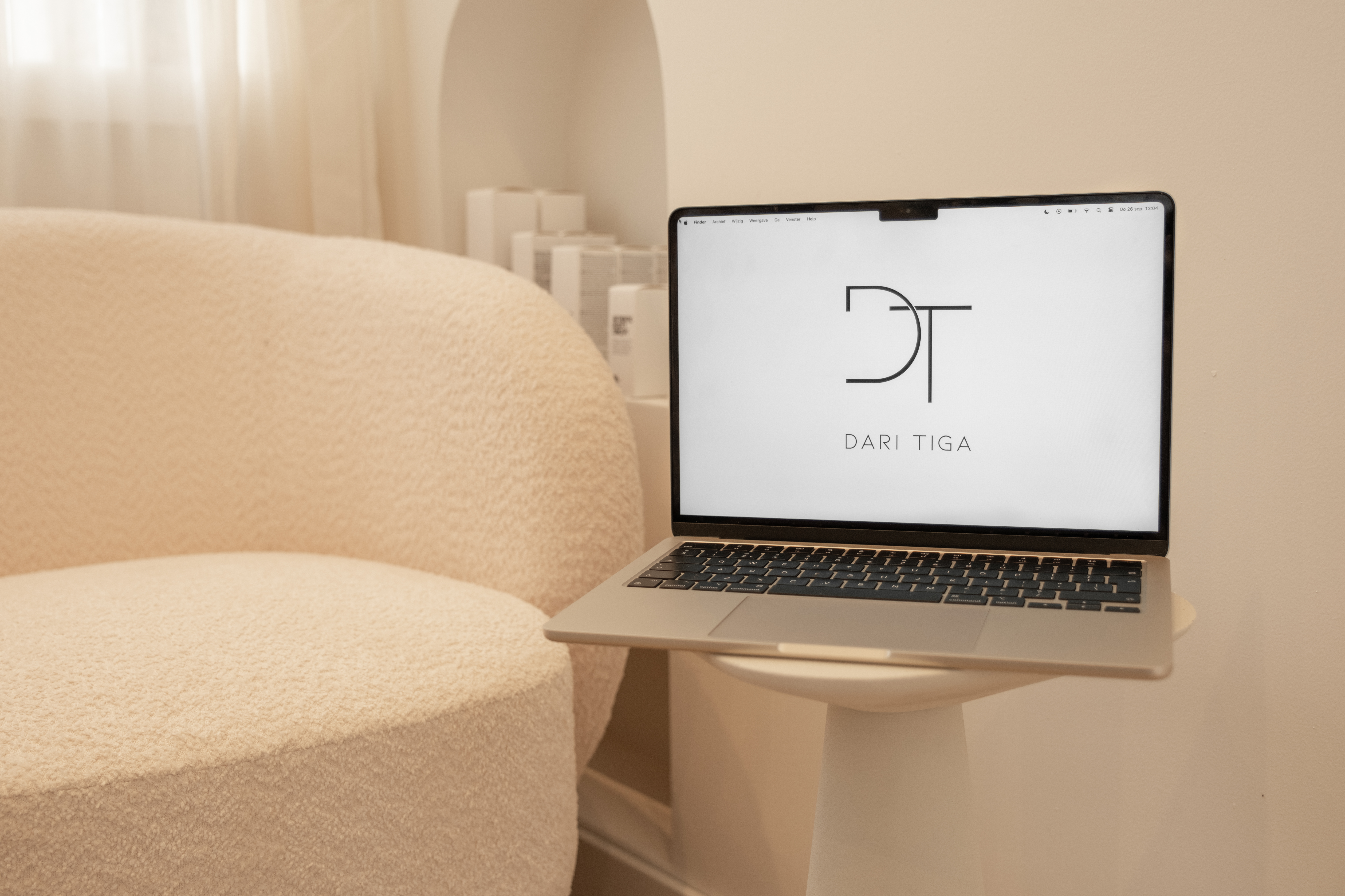 MacBook met het minimalistische Dari Tiga logo op het scherm, geplaatst op een wit tafeltje naast een bouclé stoel in Putten