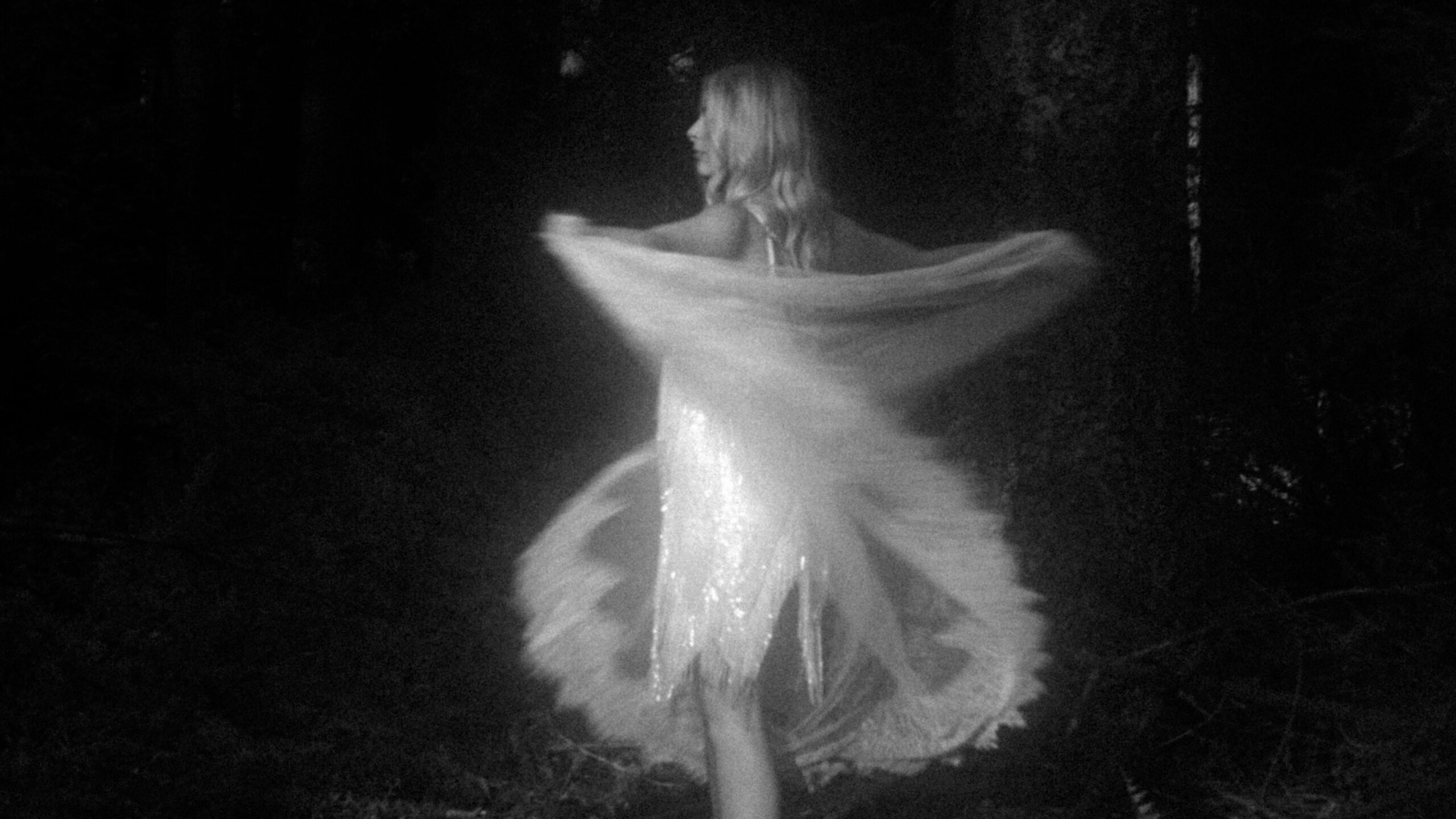 Artistieke zwart-witfoto van model Rosalie die dynamisch beweegt in een glinsterende jurk tijdens een shoot in het bos bij Ermelo voor Ivory & Woods