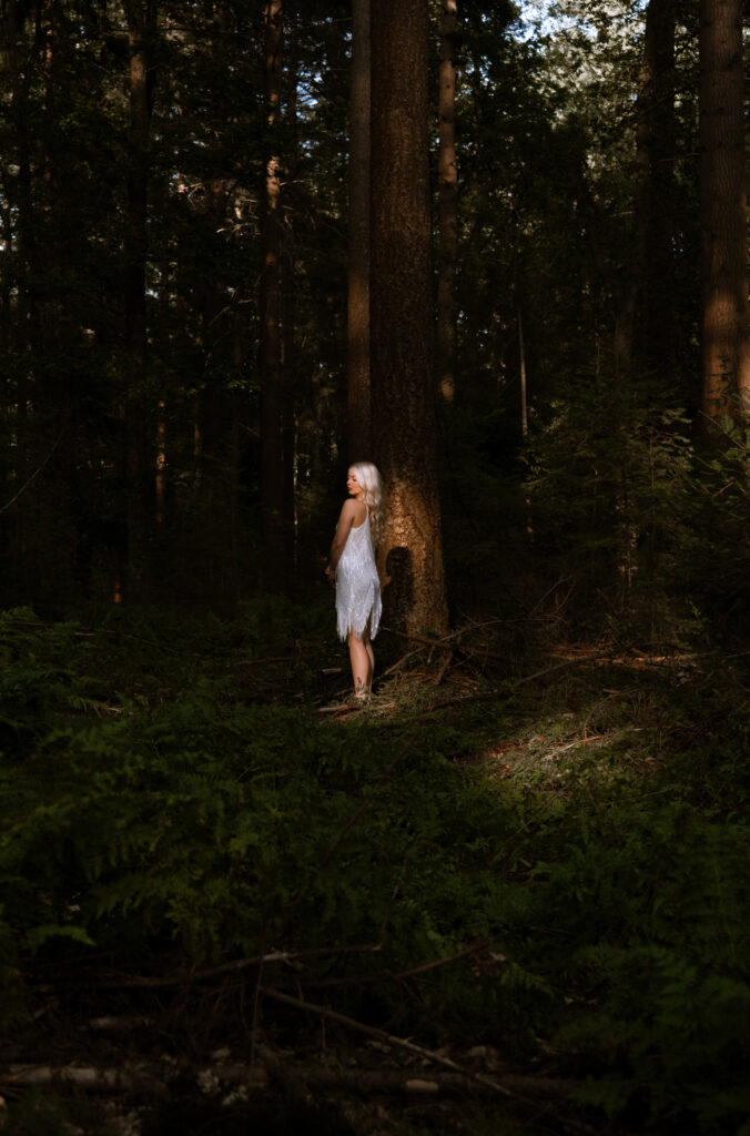 Model Rosalie in een witte jurk staand in het sfeervolle zonlicht tussen de bomen in het bos bij Ermelo voor Ivory & Woods