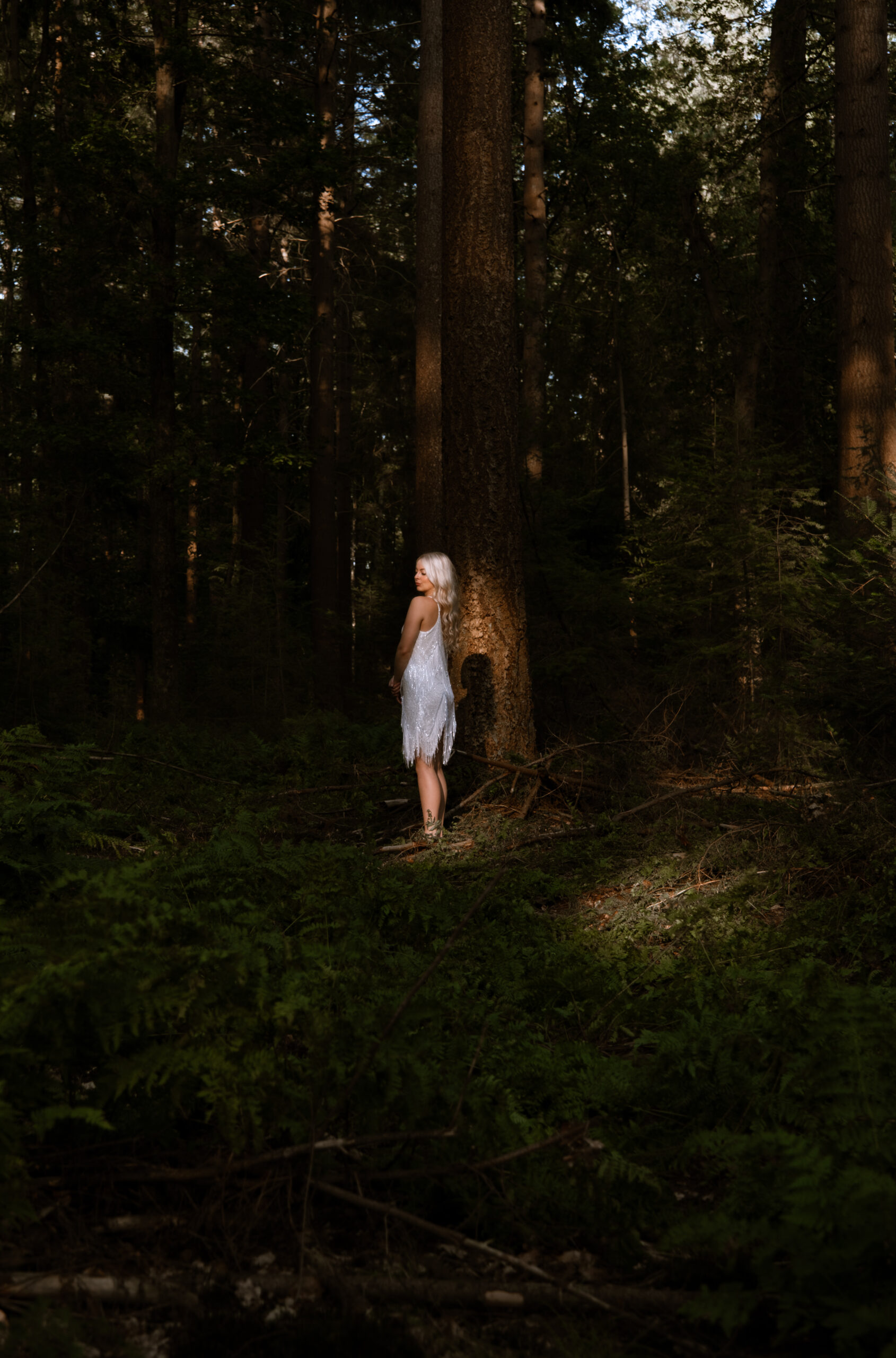 Model Rosalie in een witte jurk staand in het sfeervolle zonlicht tussen de bomen in het bos bij Ermelo voor Ivory & Woods