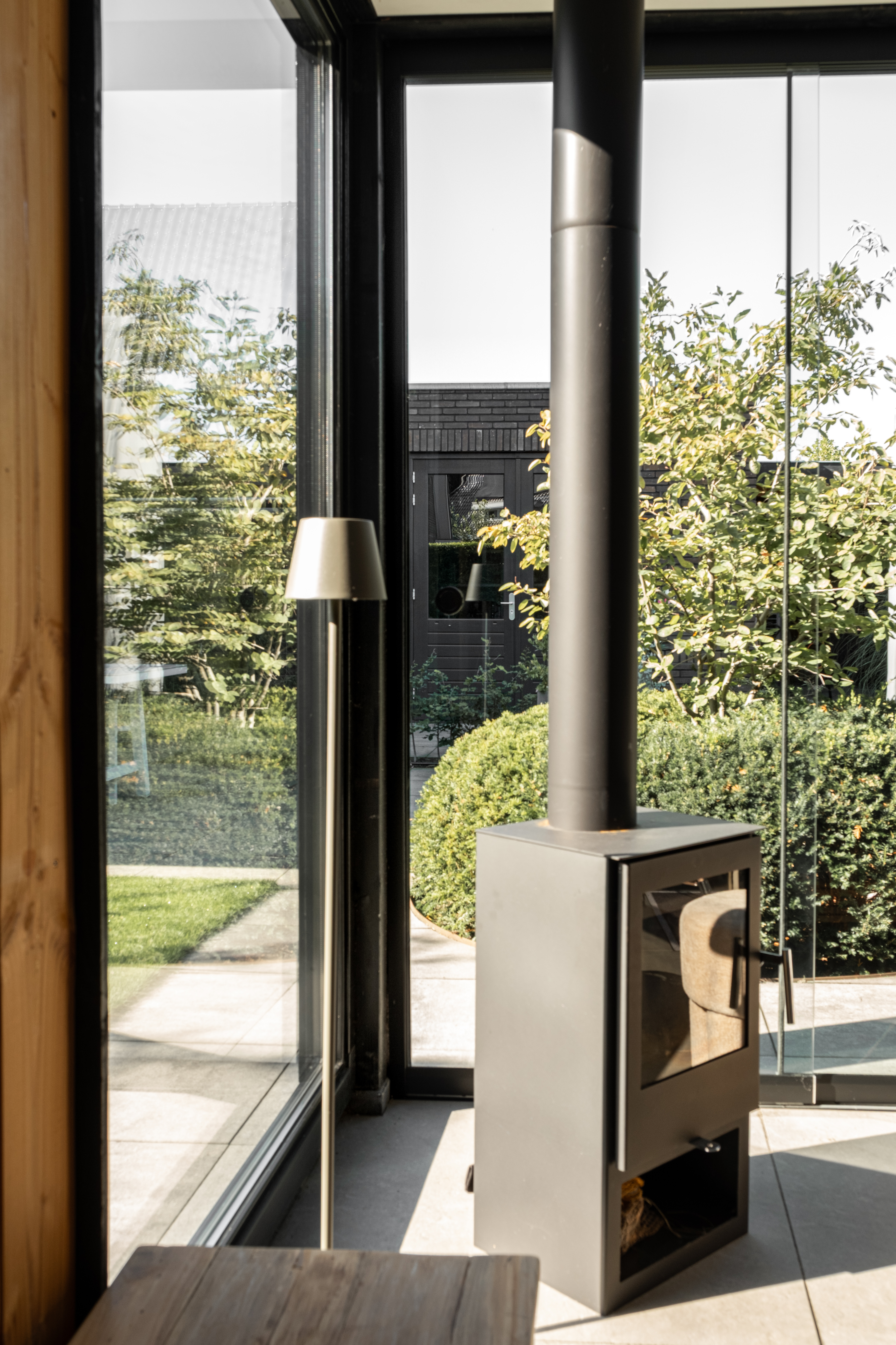 Sfeervolle hoek in een houten tuinkamer in Lunteren door BG Buitenverblijven met een vrijstaande zwarte houtkachel voor een grote glazen pui