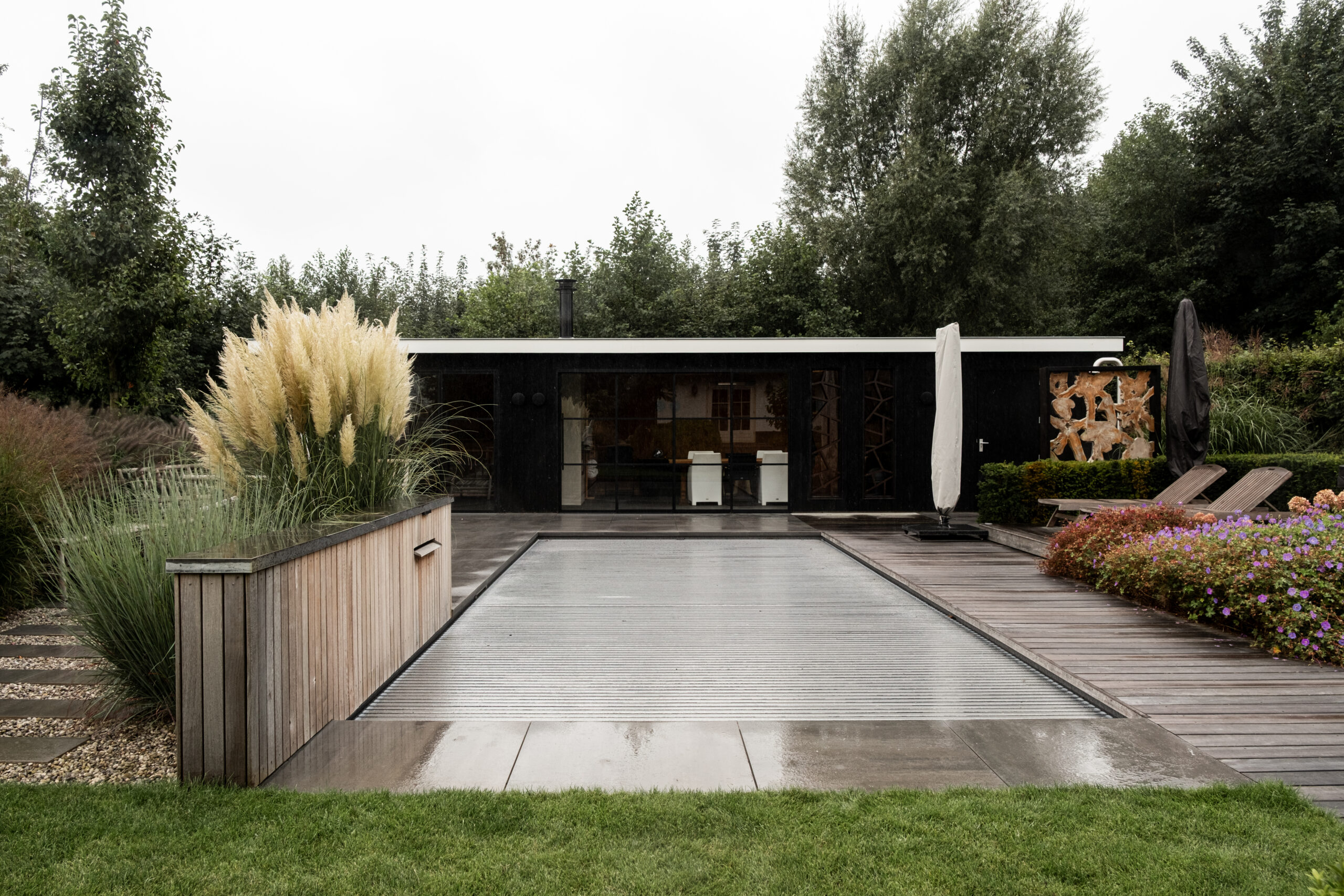 Recht overzicht van het zwarte poolhouse in Nijkerk door BG Buitenverblijven met afgedekt zwembad, houten vlonderterras en pampasgras