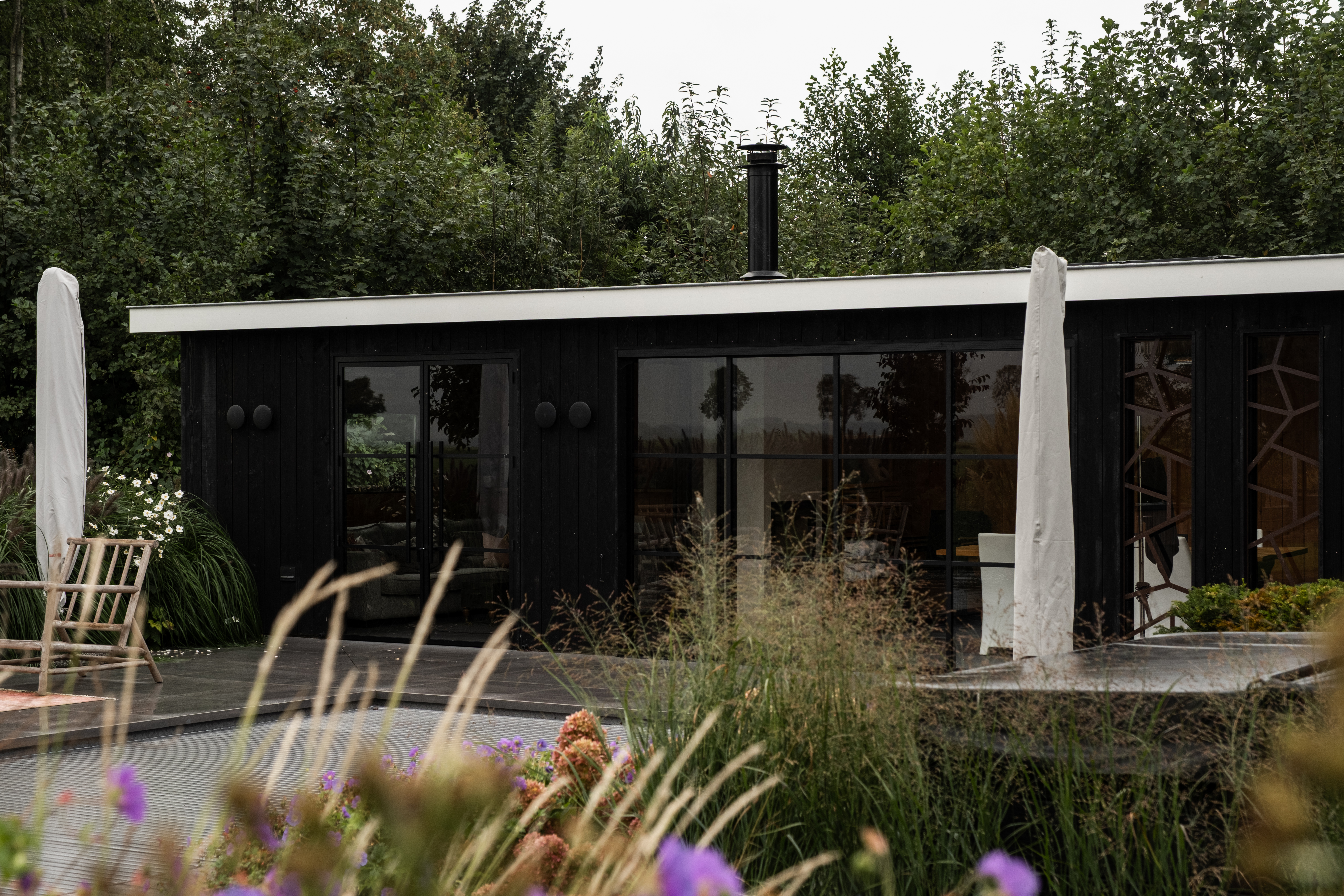 Aanzicht van het zwarte poolhouse in Nijkerk door BG Buitenverblijven met stalen pui, houten terras en zwembad omgeven door groen