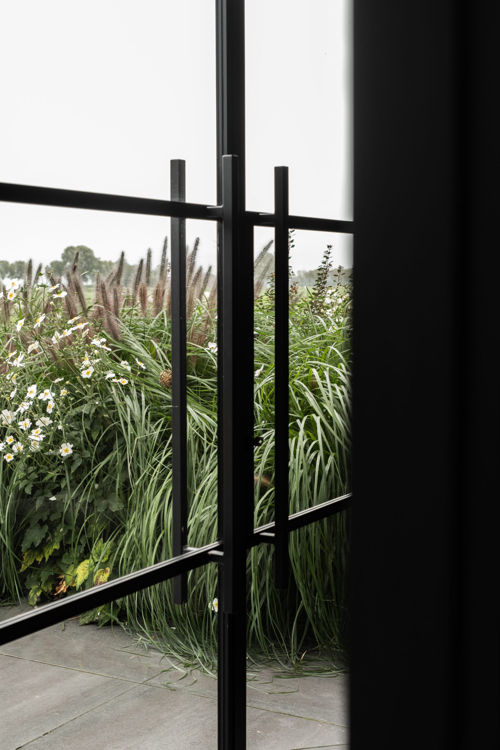 Detailopname van een minimalistische zwarte stalen deurgreep en stalen kozijn van het poolhouse in Nijkerk door BG Buitenverblijven met zicht op bloeiende siergrassen in de tuin
