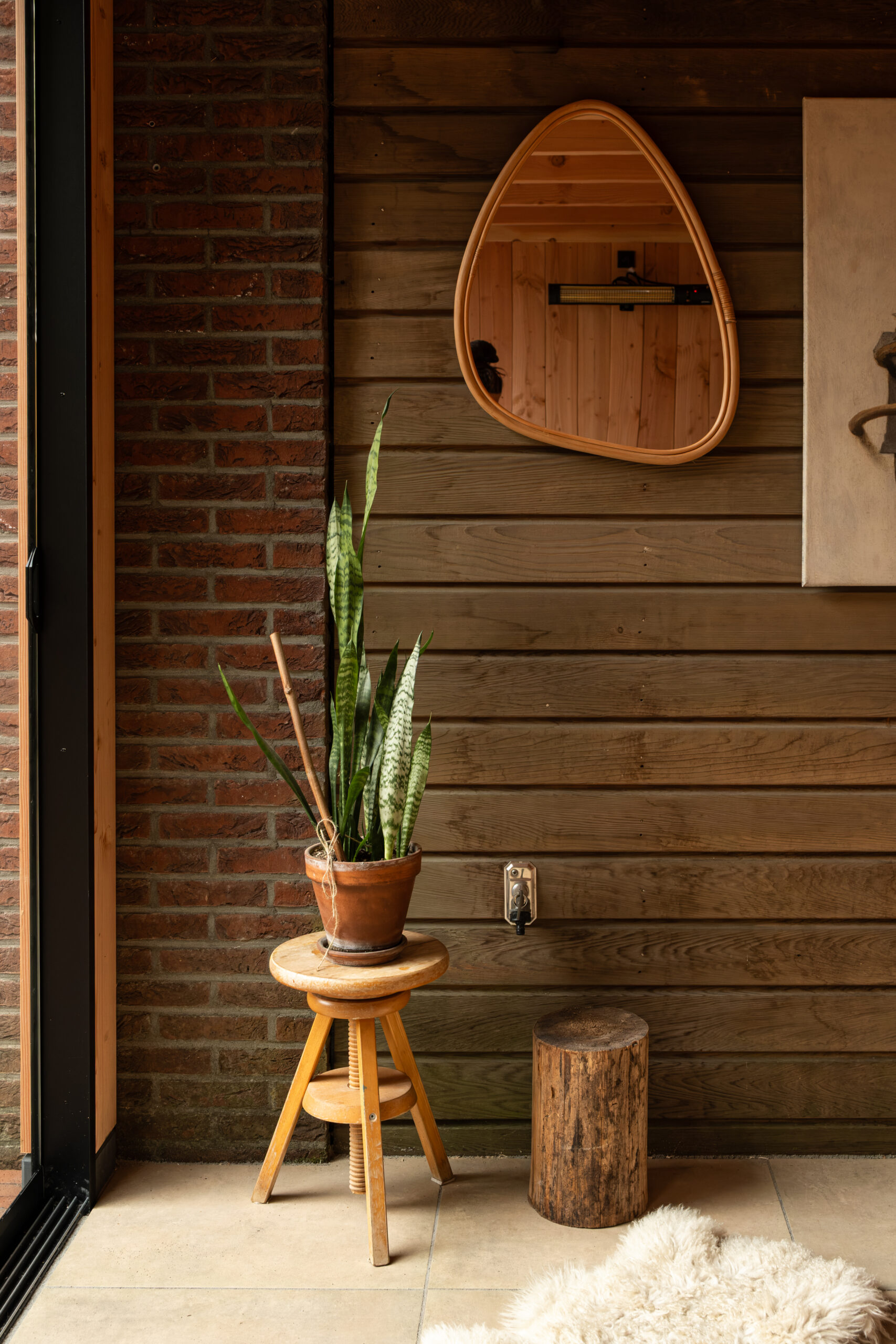 Detailopname van een houten veranda interieur in Ede met een organisch gevormde spiegel op een houten wand, een krukje met plant en een bakstenen muur door BG Buitenverblijven