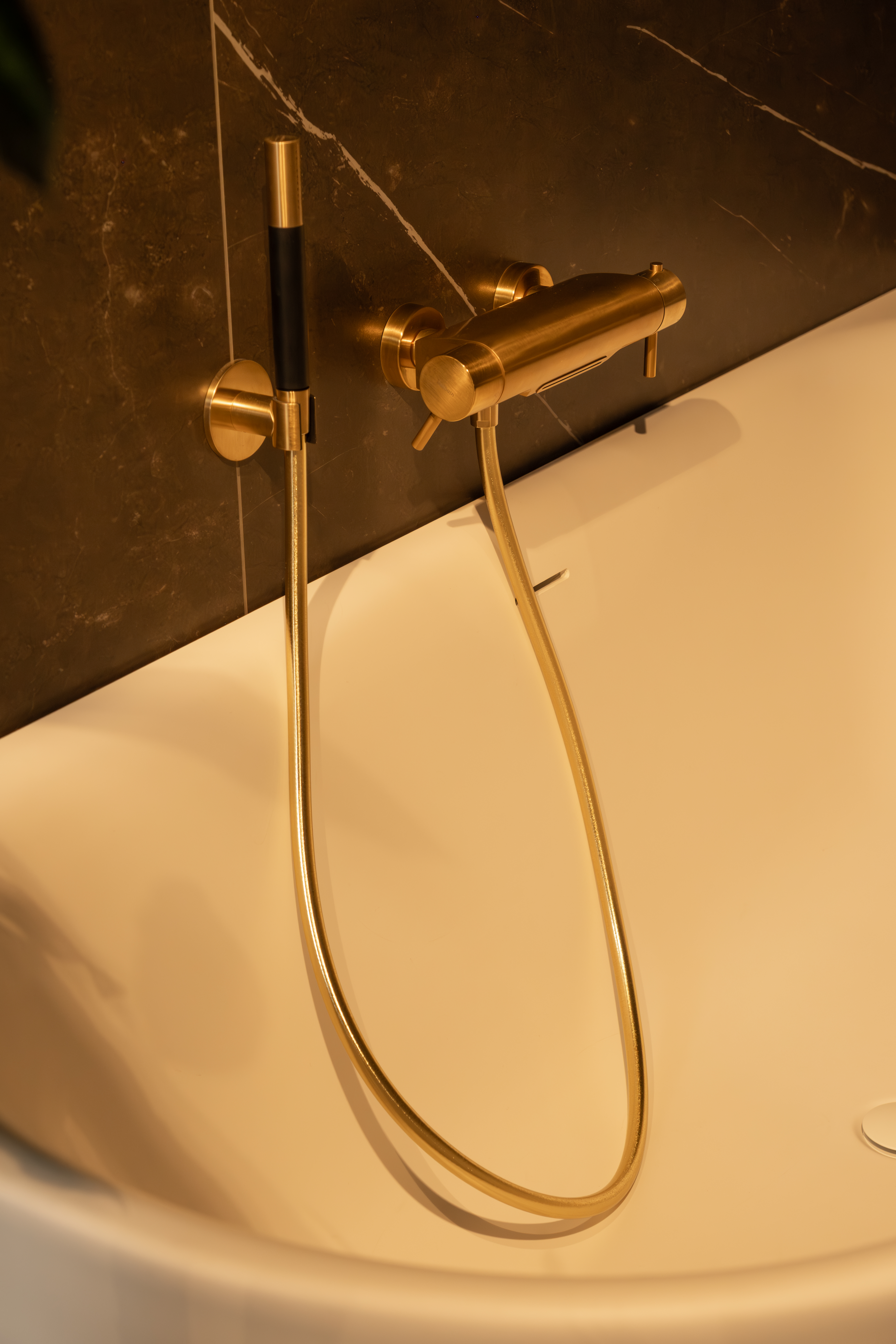 Gouden inbouwkraan en handdouche bij een wit ligbad tegen een donkere marmeren wand in de showroom van Berends Badkamers Woerden