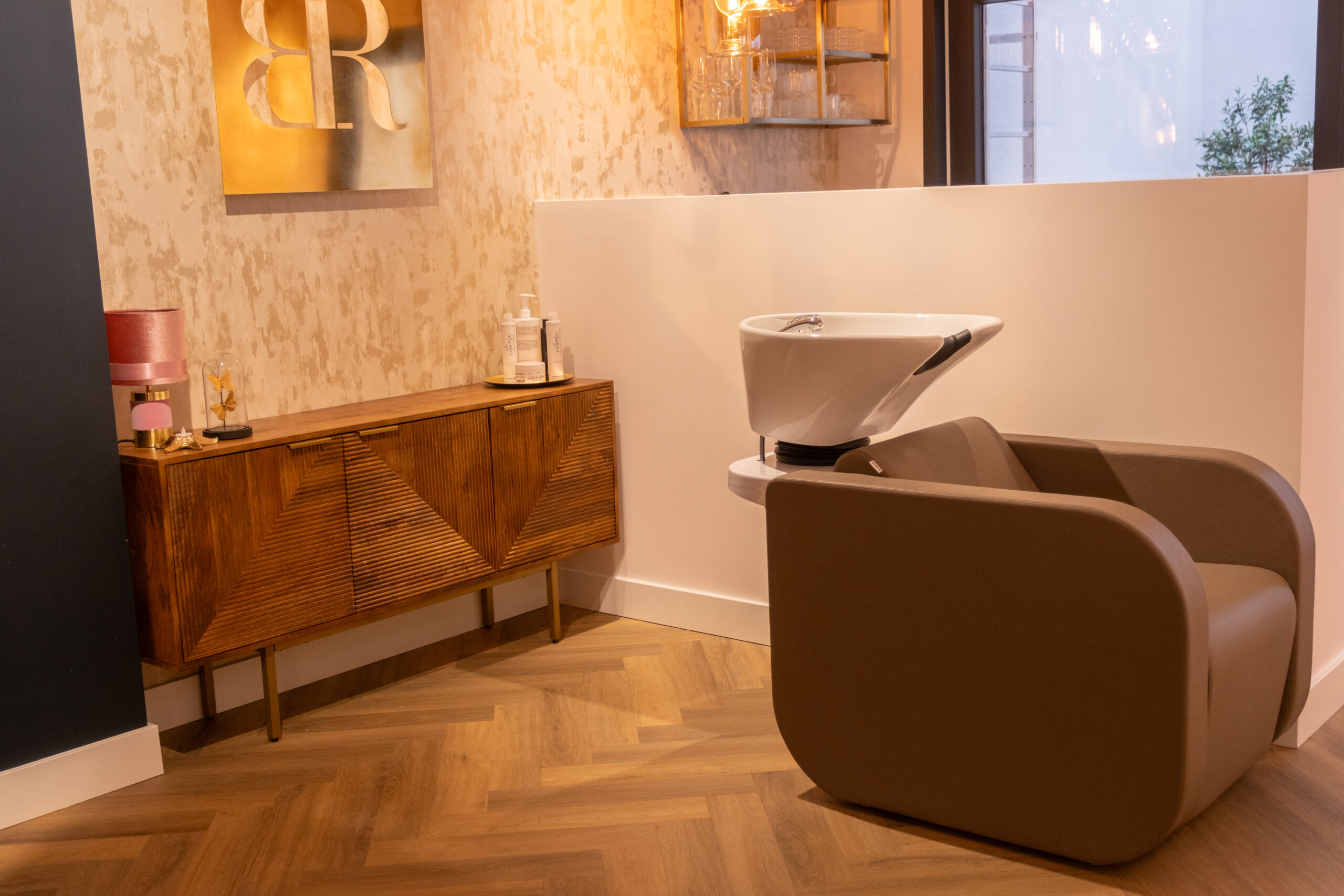 Luxe wasunit met lederen stoel en een houten dressoir met gouden wanddecoratie bij Beauty by Roos Antwerpen