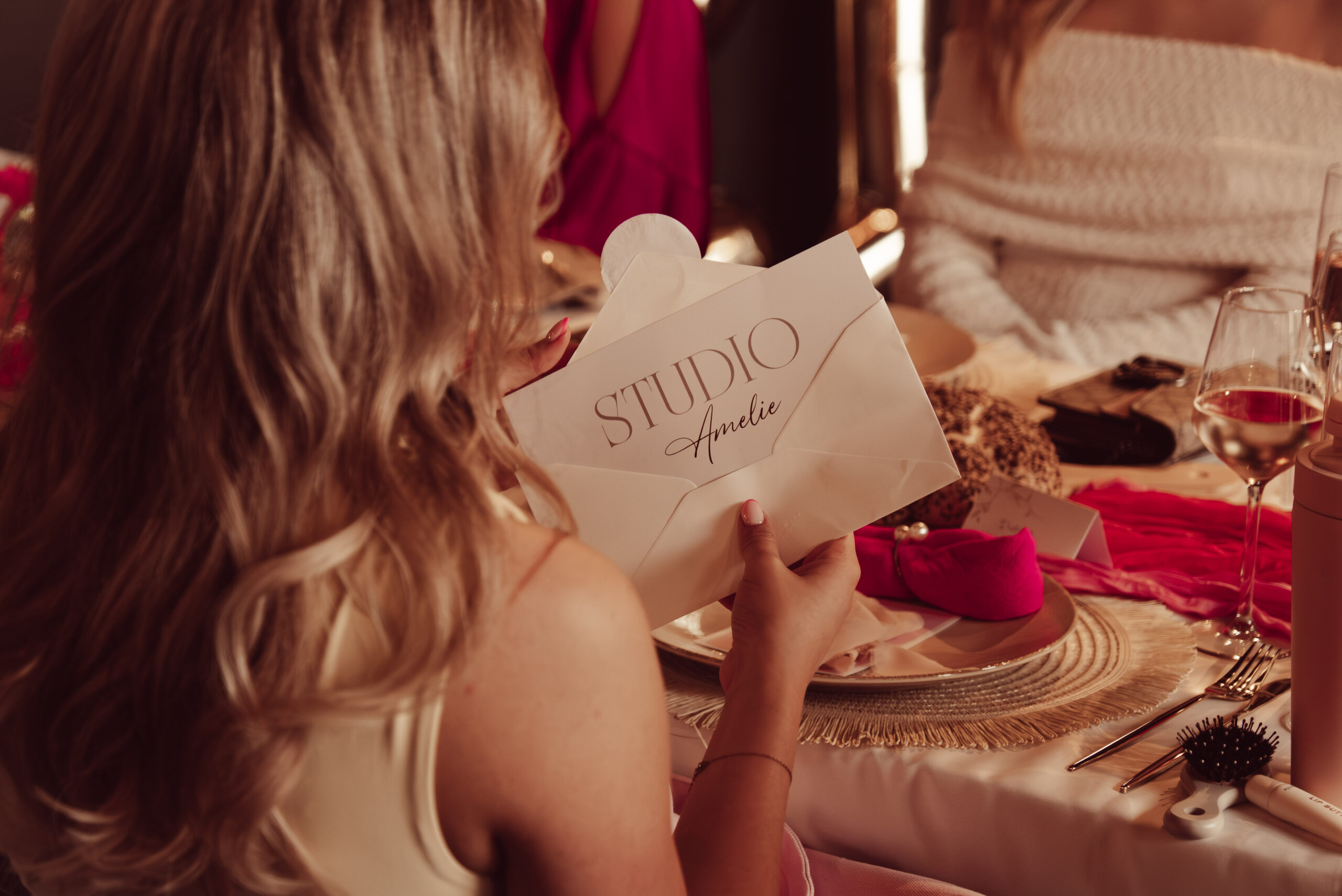 Blonde vrouw houdt een witte enveloppe met de opdruk Studio Amelie vast aan een luxe gedekte feesttafel met roze accenten
