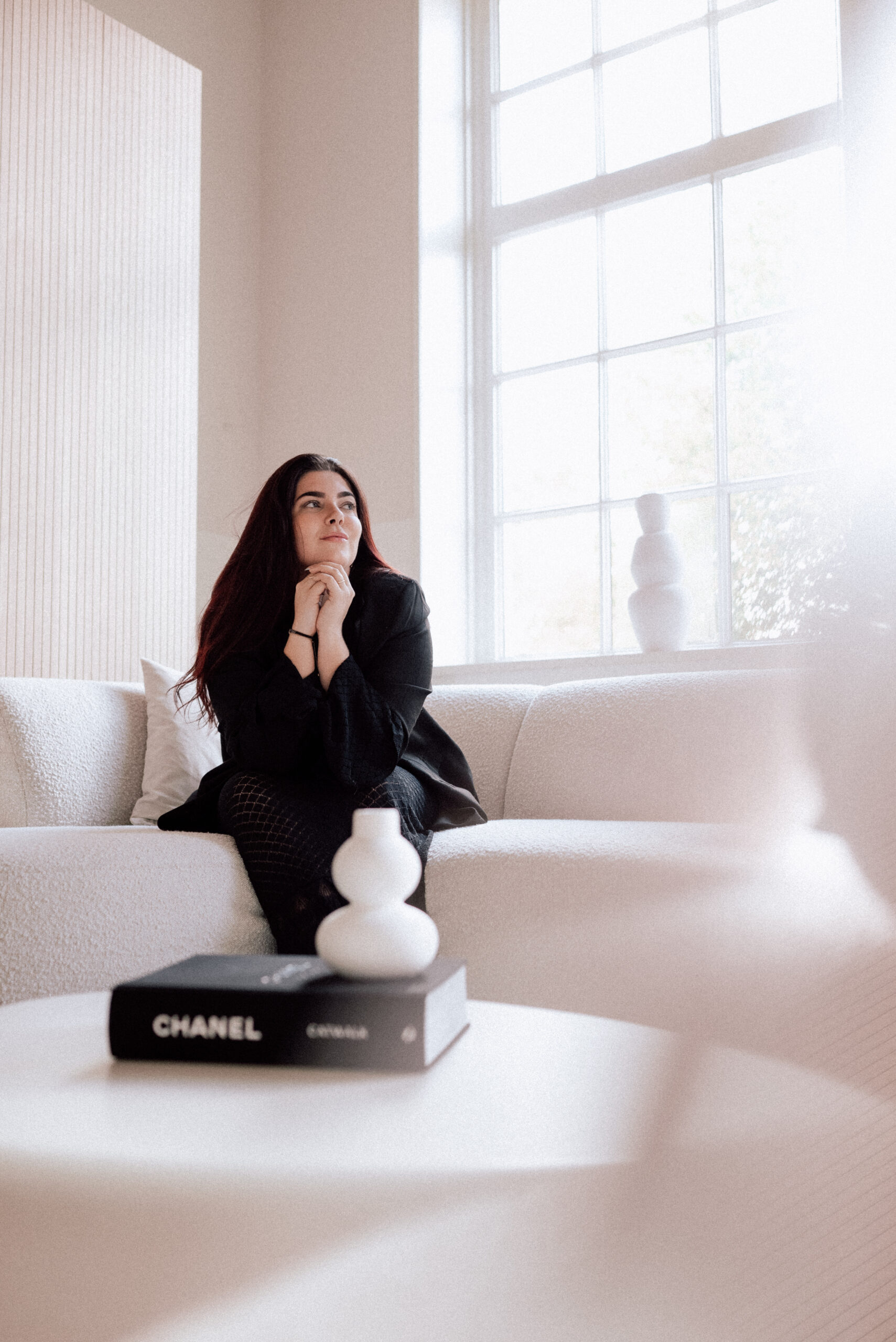 Judith Schouten zit peinzend op een witte bouclé bank voor een groot raam, met een Chanel-boek en een vaas op de voorgrond.